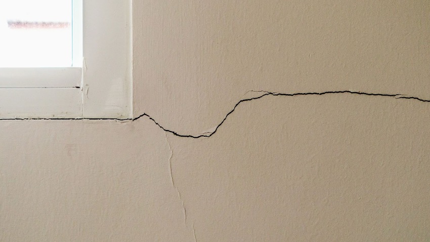 Uma rachadura na parede horizontal corre ao longo de uma parede interna esbranquiçada, começando no canto inferior da moldura de uma janela.
