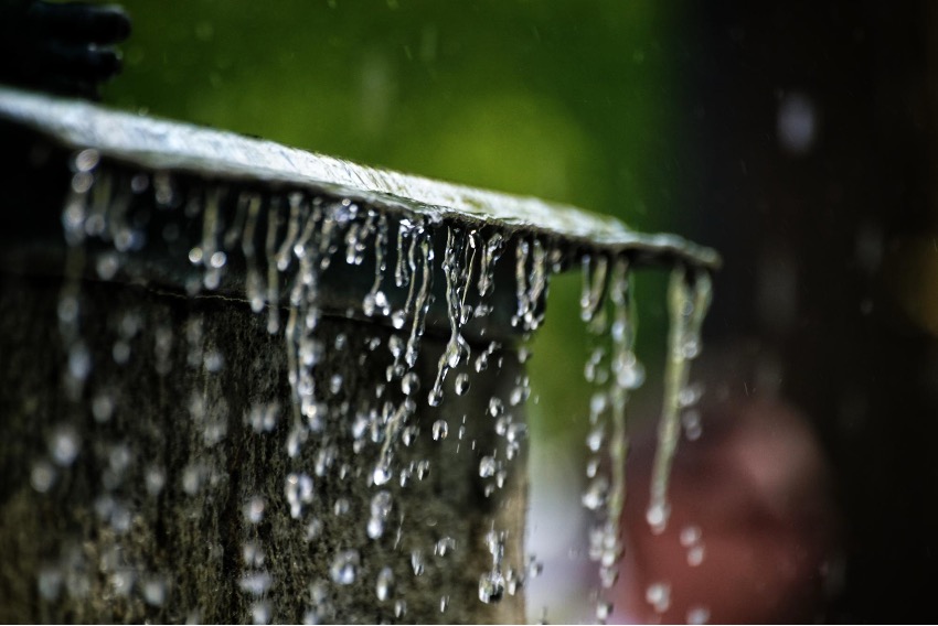 A água da chuva cai em cascata sobre uma superfície inclinada, com gotas capturadas no ar contra um fundo verde e escuro desfocado — uma representação perfeita da captação de água da chuva na natureza.