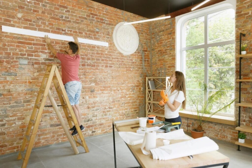 Duas pessoas trabalhando em uma reforma com escada em uma sala com parede de tijolos.