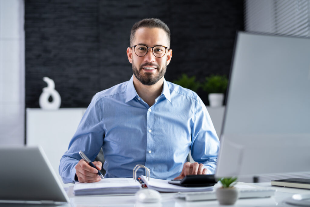 Um empresário sorridente sentado à sua mesa em frente a um computador, trabalhando no planejamento financeiro.