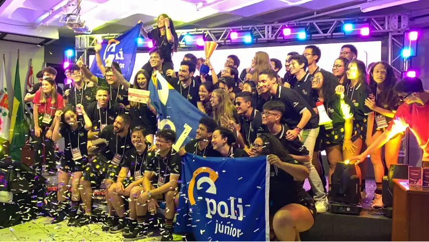 Grupo de jovens da poli junior exultantes segurando bandeiras e troféu comemorando em palco com confetes em evento da Empresa Júnior.