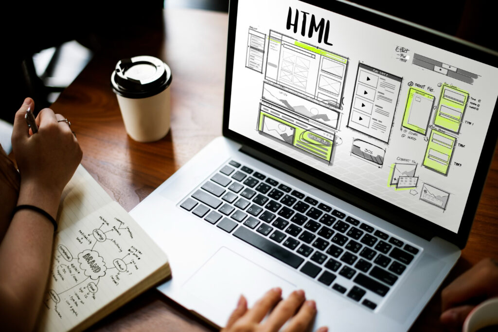 Duas pessoas colaborando em web design; um faz anotaçõe enquanto o outro usa um laptop exibindo um layout HTML. Uma xícara de café está sobre a mesa.