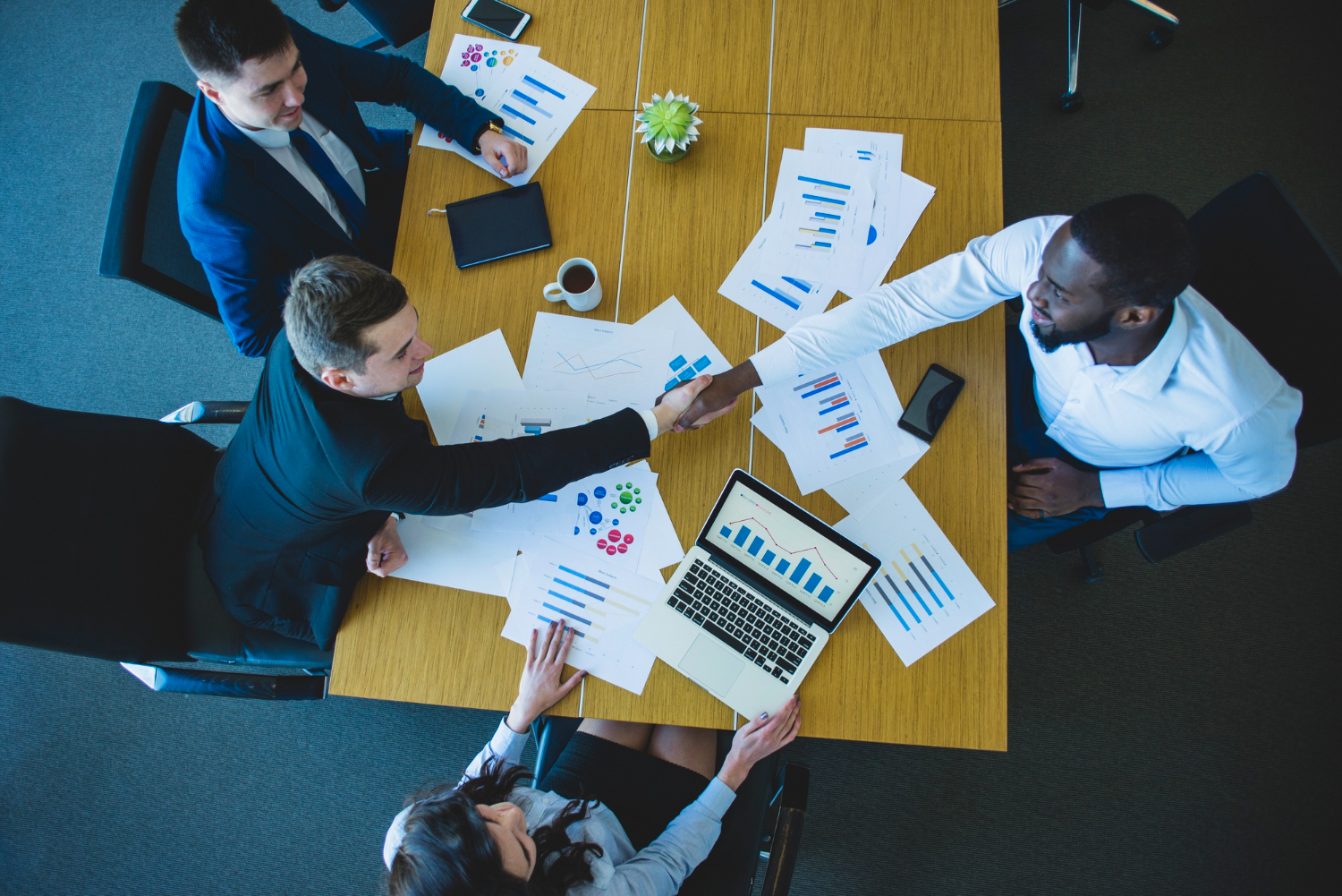 Quatro pessoas em uma reunião de negócios, sentadas ao redor de uma mesa com gráficos e tabelas. Dois participantes homens apertam as mãos, indicando um acordo. A cena reflete uma consultoria em gestão empresarial para uma empresa