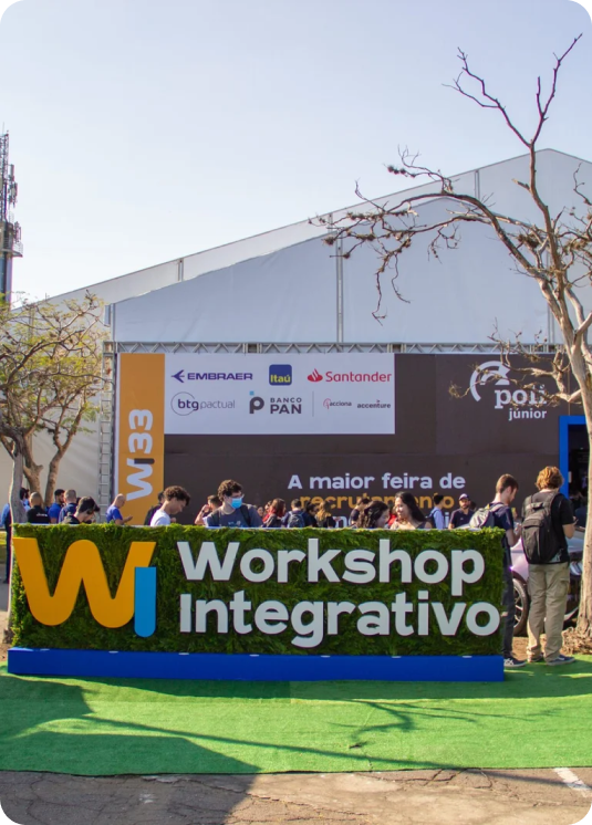 A cena ao ar livre apresenta uma grande placa com os dizeres "Workshop Integrativo", cercada por vários logotipos de patrocinadores. As pessoas estão reunidas ao redor, com árvores ao fundo e uma tenda branca visível.