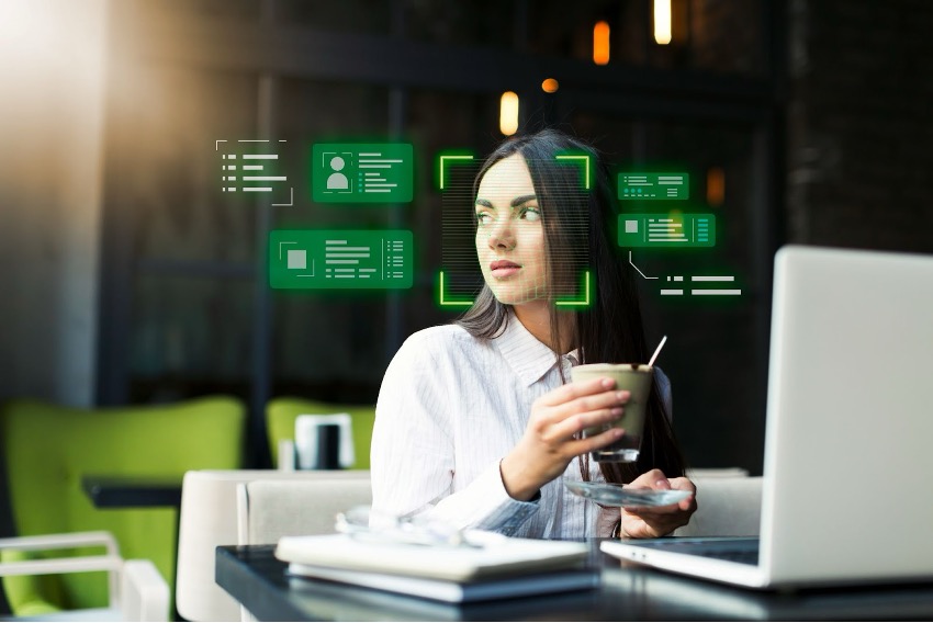 Uma mulher está sentada à mesa segurando uma xícara enquanto olha para o lado. Um laptop está na frente dela, e gráficos de interface digital verde, talvez exibindo aplicações de Inteligência Artificial, estão sobrepostos perto de seu rosto.