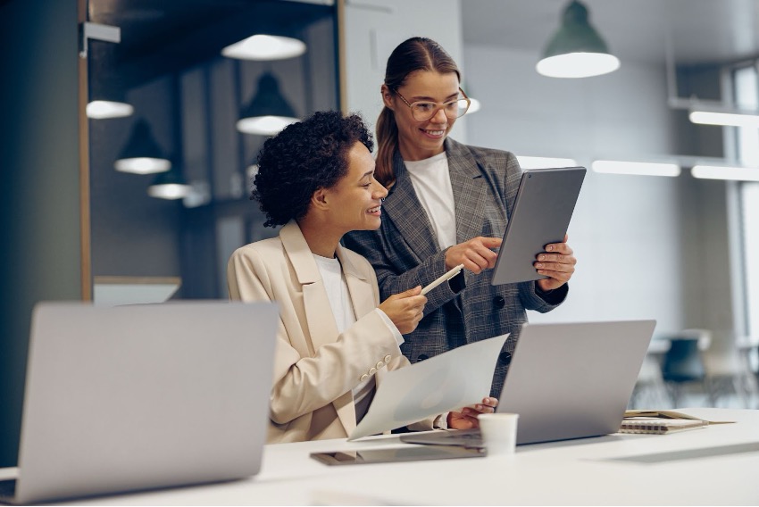 Dois profissionais em um ambiente de escritório, um segurando um tablet com o "Sistema SAP: Guia Completo" e apontando, enquanto o outro sorri e segura um documento. Laptops são visíveis na mesa.