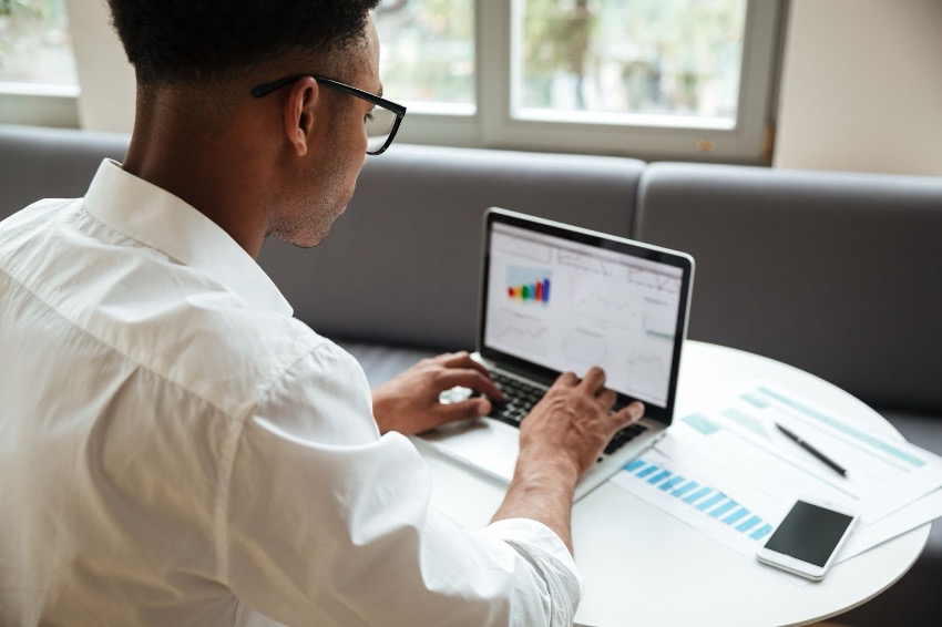 Um homem de camisa branca trabalha em um laptop exibindo gráficos e tabelas do seu Power BI Dashboard de Vendas, sentado a uma mesa com papéis e um smartphone.