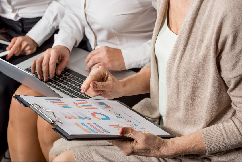 Pessoas em trajes profissionais da Poli Júnior estão revisando gráficos e dados em uma prancheta e um laptop, concentrando-se em transformar resultados por meio de pesquisas de mercado detalhadas.