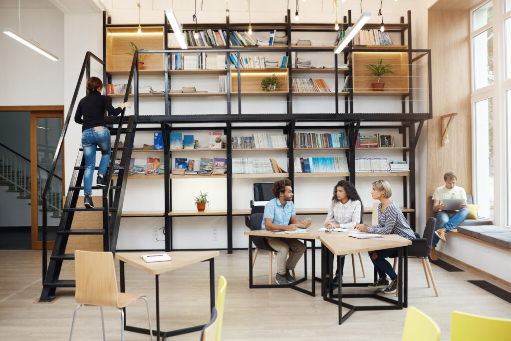 Em uma empresa moderna com design de interiores elegante, uma pessoa sobe uma escada para alcançar livros, enquanto outras três discutem em uma mesa.