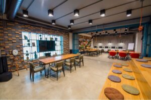 Um espaço de coworking moderno com mesas longas de madeira, cadeiras e decoração estilosa em prateleiras de parede. O design de 2025 apresenta uma fileira de estações de computador, complementadas por piso de concreto e iluminação superior elegante.