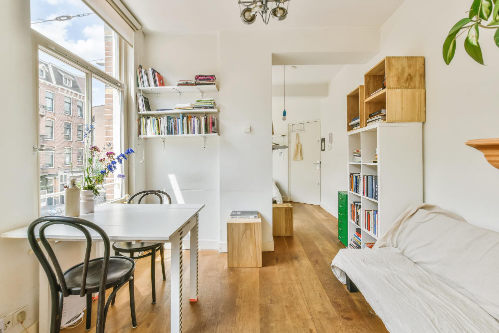 Apartamento compacto e iluminado com mesa de jantar, cadeiras, estantes e piso de madeira, perfeito para quem quer decorar casa pequena e aproveita cada metro, com um sofá aconchegante perto de janelas grandes e paredes brancas para uma sensação arejada.