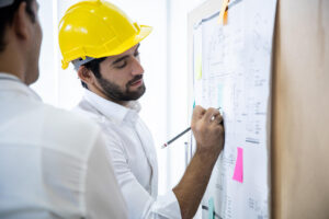 Engenheiro com capacete amarelo analisando e marcando planta arquitetônica em painel com post-its coloridos, durante o planejamento de obras na construção civil.