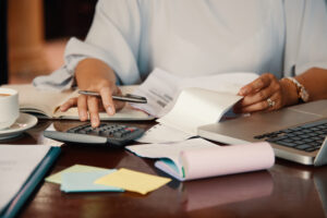 Pessoa usando uma calculadora e segurando papéis em uma mesa com um laptop, notebook, xícara de café, notas adesivas e documentos sobre modelo de faturamento e previsão de faturamento.