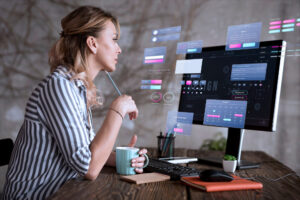 Uma mulher está sentada em uma mesa segurando uma caneca, olhando para um monitor de computador que exibe várias janelas de interface UX holográfica, com foco em aprimorar a experiência do usuário.