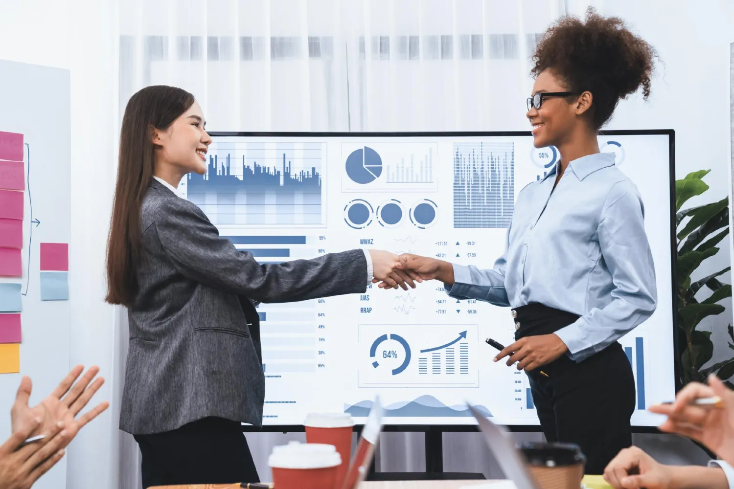 Duas empresárias apertam as mãos em uma sala de reunião de escritório, em pé diante de uma tela exibindo vários gráficos e tabelas, sinalizando uma reestruturação comercial bem-sucedida.