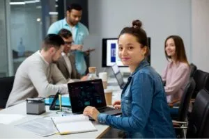 Uma mulher em uma jaqueta jeans está sentada em uma mesa com um laptop exibindo gráficos,discussão no fundo do ambiente de escritório moderno.