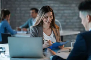 Representação de uma consultoria bpm, uma mulher sorridente segurando uma prancheta está sentada em uma mesa com um laptop, discutindo com um homem. Duas pessoas colaboram ao fundo.