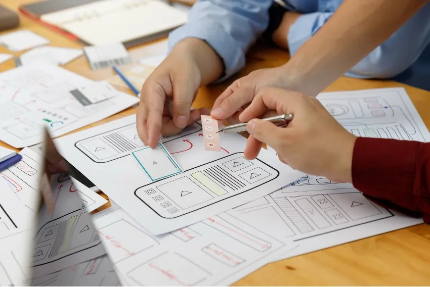 Mãos esboçam wireframes de aplicativos , usando canetas e papel espalhados pela mesa. Esses rascunhos servem como exemplos cruciais no desenvolvimento de estratégias que melhoram o ROI do processo de Product Discovery.