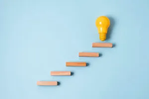 Cinco blocos de madeira formam degraus sobre um fundo azul, levando até uma lâmpada amarela no topo, simbolizando como aplicar na prática a inovação incremental rumo a novas ideias.