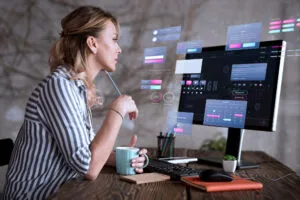 Uma mulher está sentada em uma mesa segurando uma caneca, olhando para um monitor de computador que exibe várias janelas de interface UX holográfica, com foco em aprimorar a experiência do usuário.